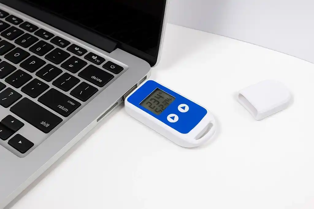 Mini termohigrómetro con registro de datos conectado al usb de un ordenador portátil encima de una mesa blanca