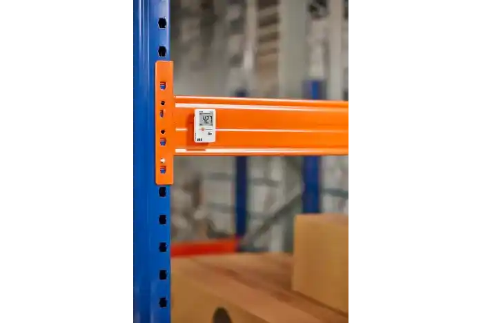 Termohigrómetro digital profesional con función de registro de datos de temperatura y humedad relativa, modelo Testo 174H en su variante en color blanco, fijado en la estructura de una estantería industrial naranja y azul de un almacén