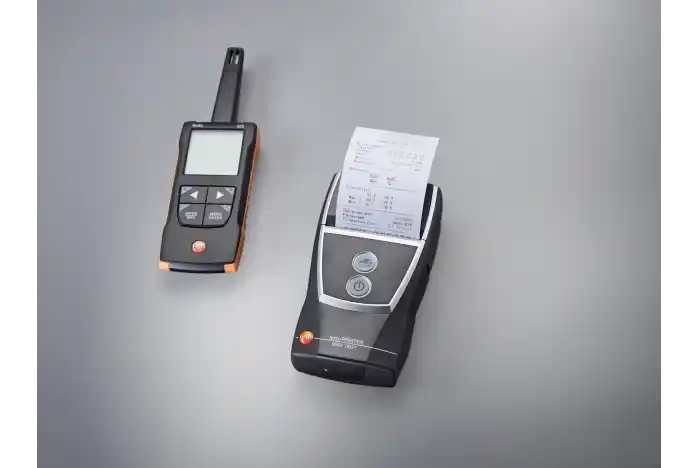 Termohigrómetro testo 625 y a su derecha una pequeña impresora testo 0554 0622 bluetooth, ambos encima de una superfície gris claro