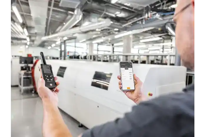 Técnico con gafas sosteniendo con la mano izquierda el termohigrómetro profesional y con la derecha un smartphone que muestra los valores del termohigrómetro portátil mediante bluetooth, en el interior de una empresa