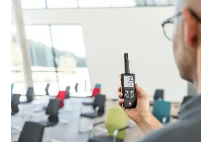 Persona con gafas sosteniendo un termohigrómetro Testo 625 con la mano derecha mientras mide los valores de temperatura humedad relativa y punto de rocío de una moderna sala de actos