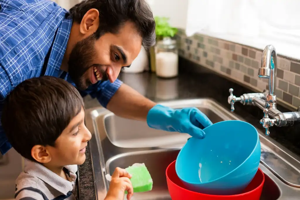 Padre sonriente con camisa a cuadros azules y guantes de limpieza, lavando platos junto a su hijo pequeño en la cocina, disfrutando del momento juntos